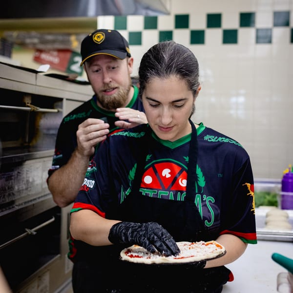 Two Mattenga's Pizzeria employees preparing a pizza with sauce and cheese in a kitchen.