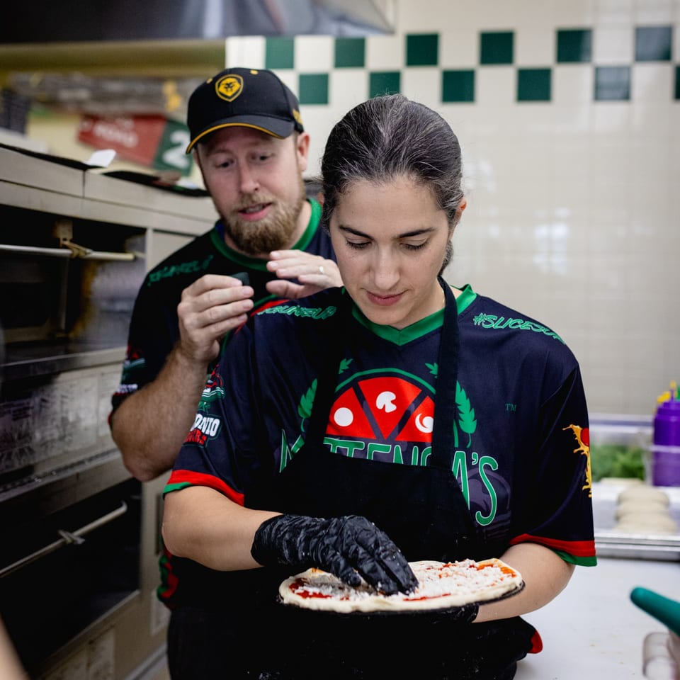 Two Mattenga's Pizzeria employees preparing a pizza with sauce and cheese in a kitchen.