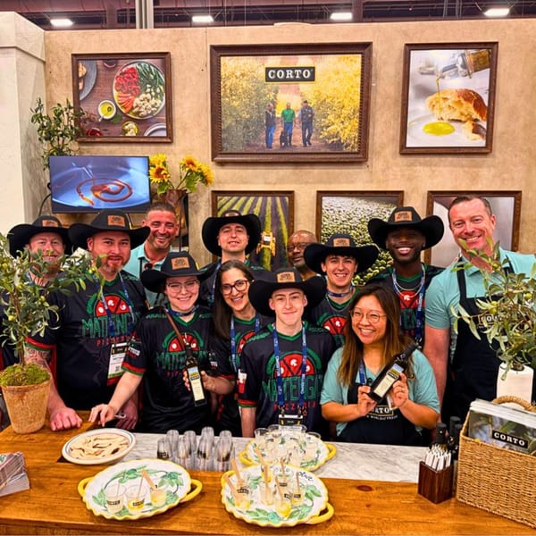 A large group of smiling people in black shirts and cowboy hats stand behind a table with food samples.