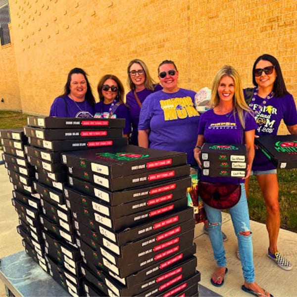 Six women in purple shirts stand beside a large stack of Mattenga's Pizzeria pizza boxes.