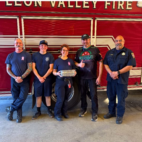 Five people, including firefighters, stand in front of a red fire truck, one holding a tray of food.