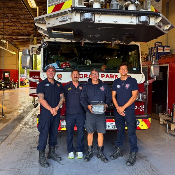 Four firefighters stand in front of a fire truck, one holding a stack of food containers.
