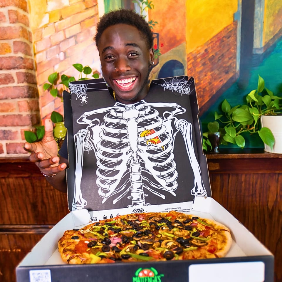 Smiling man in a skeleton pizza box costume holding a Mattenga's Pizzeria pizza.
