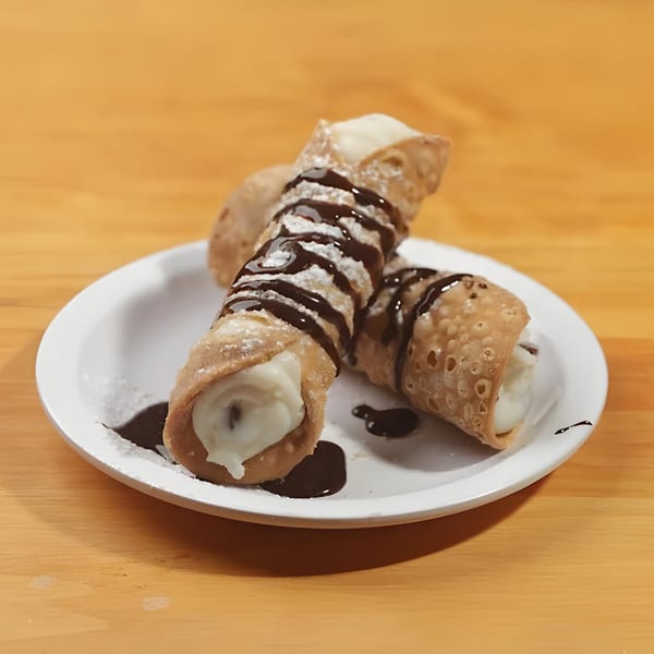 Two cannolis with powdered sugar and chocolate drizzle on a white plate at Mattenga's Pizzeria.
