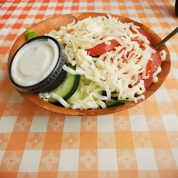 A fresh side salad with tomatoes, cucumbers, and shredded cheese, with a side of ranch dressing.