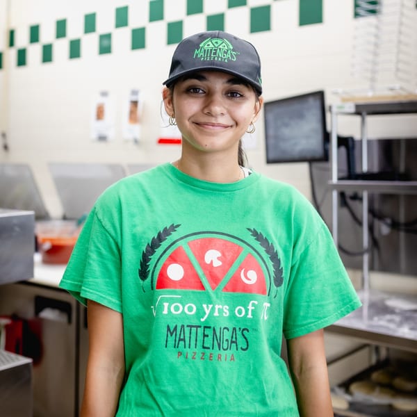Smiling Mattenga's Pizzeria employee in a green shirt and branded hat stands in the kitchen.
