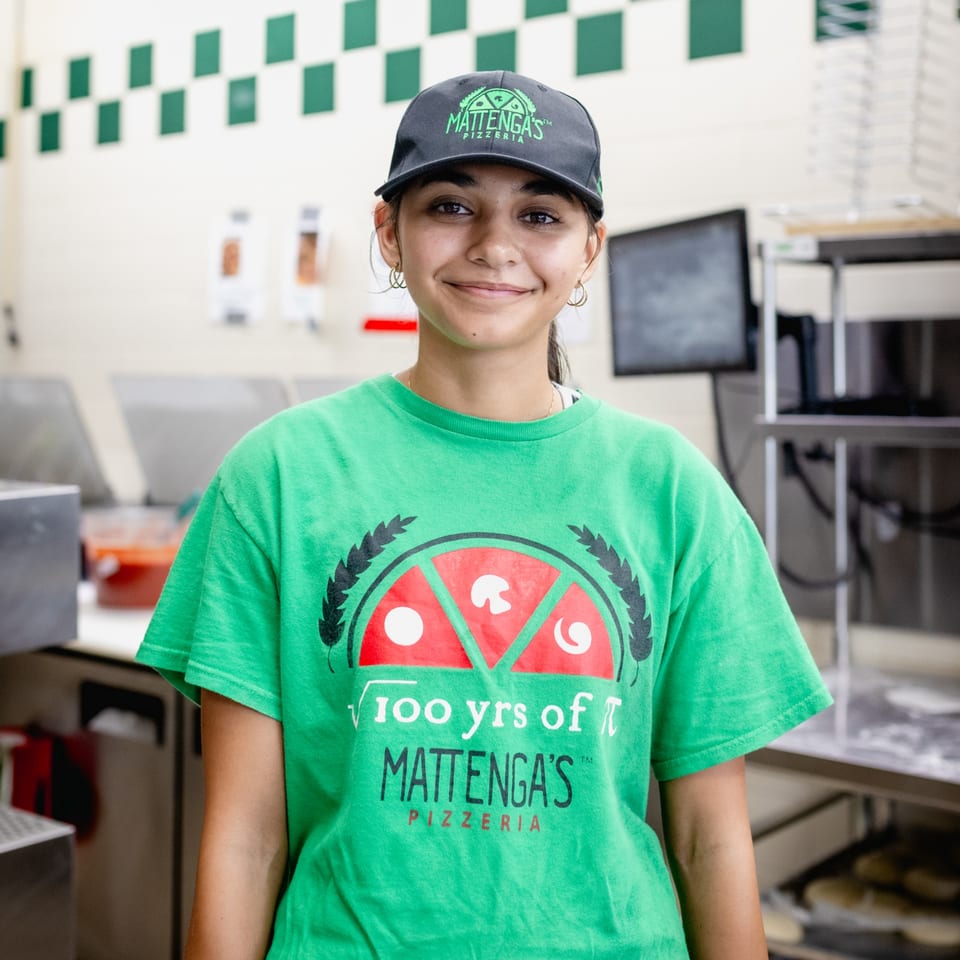 Smiling Mattenga's Pizzeria employee in a green shirt and branded hat stands in the kitchen.