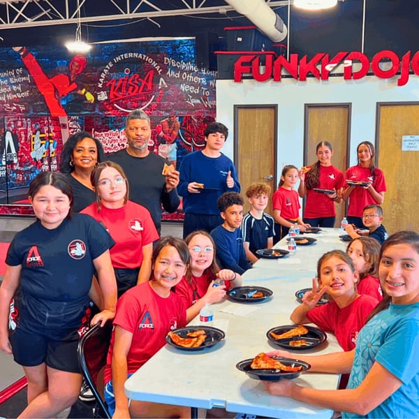 A group of children and adults smiling while eating pizza at a long table in a Funky Dojo.