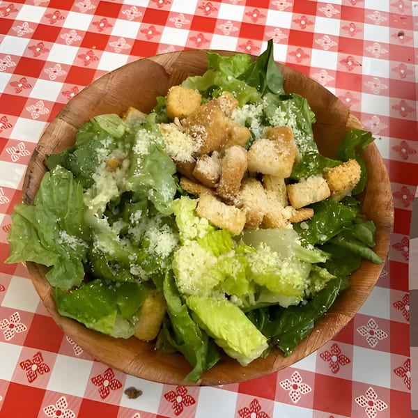 Caesar salad with romaine lettuce, croutons, and parmesan cheese in a wooden bowl on a red and white checkered tablecloth.