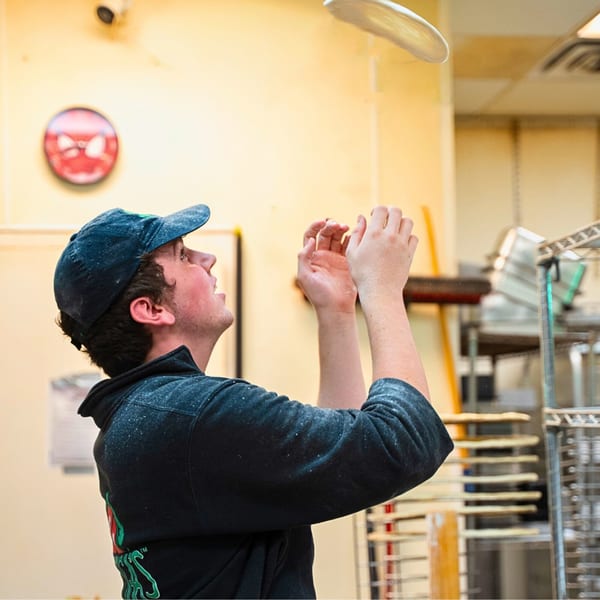 A pizza maker in a blue cap and hoodie tosses pizza dough into the air.