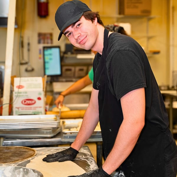 A young man in a black cap and apron smiles while preparing dough in a commercial kitchen.