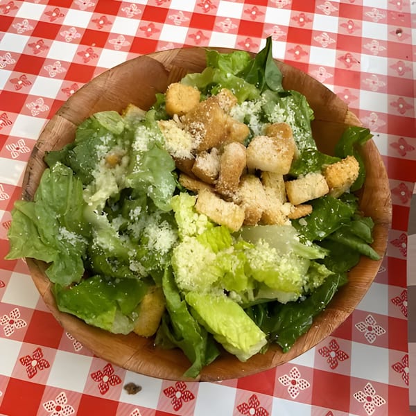 A wooden bowl of fresh Caesar salad with croutons and parmesan cheese on a red and white checkered tablecloth.