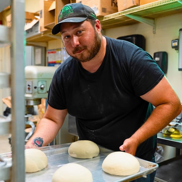 Baker in a black cap and shirt holding a tray of dough balls in a commercial kitchen.