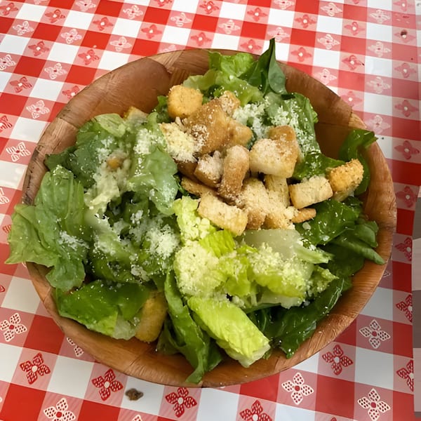 A wooden bowl of Caesar salad with croutons and parmesan cheese on a red and white checkered tablecloth.