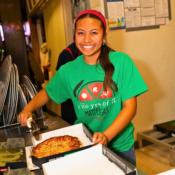 A smiling Matteyga's Pizzeria employee in a green shirt cutting a pizza in a box.