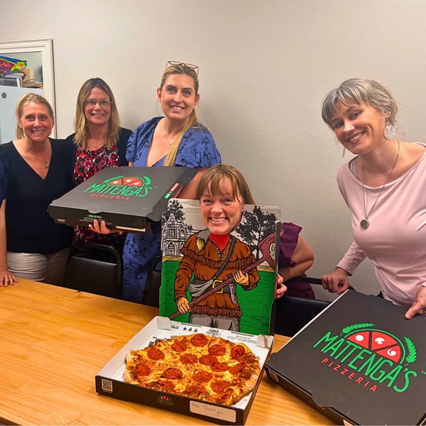 Five smiling women with Mattinga's Pizzeria boxes and a pepperoni pizza on a wooden table.
