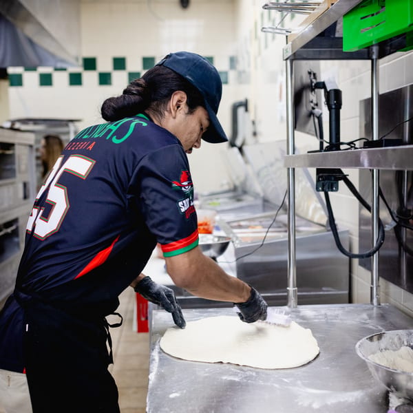 A Mattenga's Pizzeria employee in a blue uniform preparing pizza dough on a stainless steel counter.