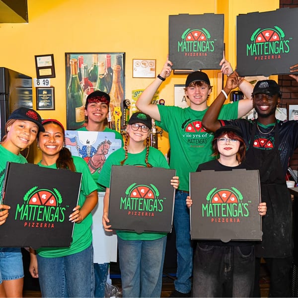 Six smiling Mattenga's Pizzeria employees in green shirts holding pizza boxes in a restaurant.
