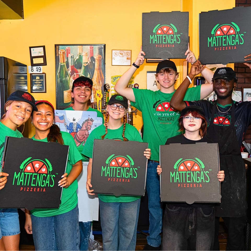 Six smiling Mattenga's Pizzeria employees in green shirts holding pizza boxes in a restaurant.