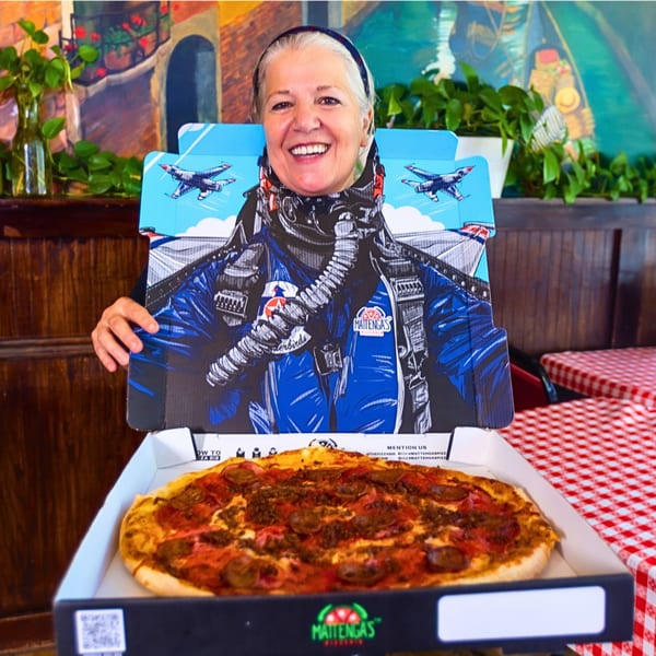 A smiling woman holds a Mattenga's Pizzeria box with a pilot cutout, revealing a meat-topped pizza.