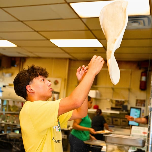 A young man in a yellow shirt expertly tosses pizza dough high into the air in a busy kitchen.