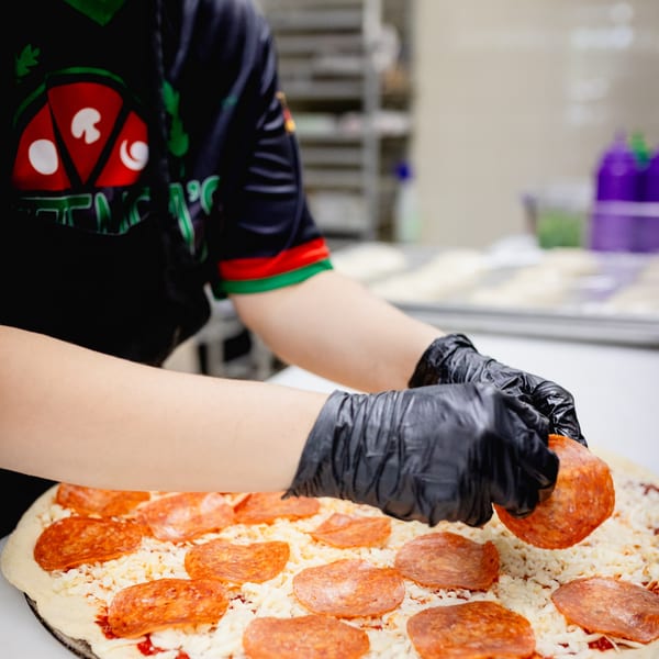 A Mattenga's Pizzeria employee in black gloves places pepperoni on a pizza with cheese and sauce.