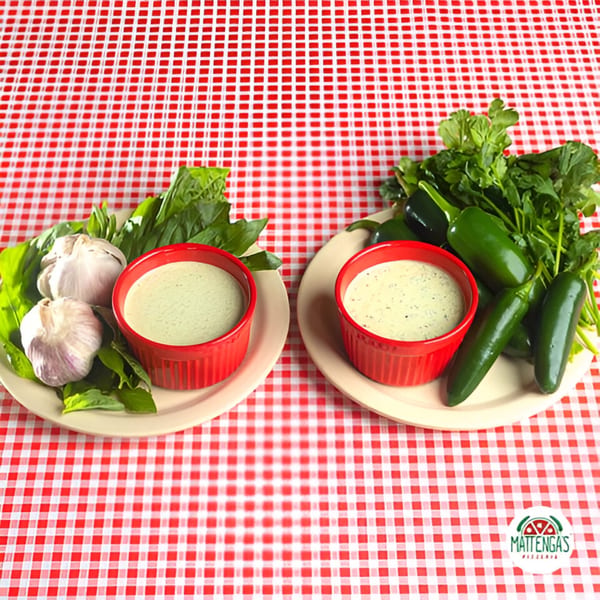 Two red ramekins of creamy sauce with garlic, basil, jalapeños, and cilantro on a red and white checkered tablecloth.