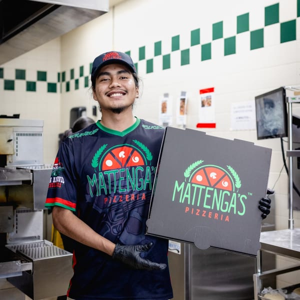 Smiling Mattenga's Pizzeria employee in uniform holding a branded pizza box in a kitchen.