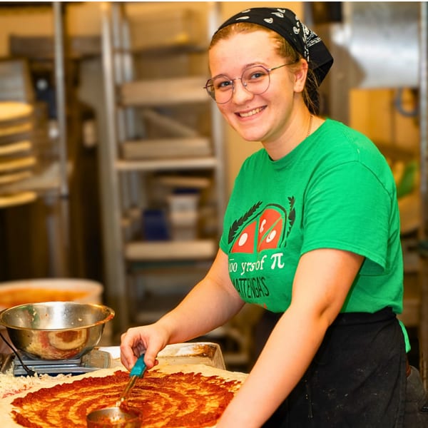 Smiling young woman in glasses and a green shirt spreading sauce on a pizza crust.