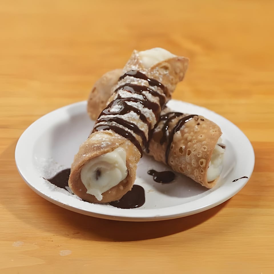 Two cannolis with powdered sugar and chocolate drizzle on a white plate at Mattenga's Pizzeria.