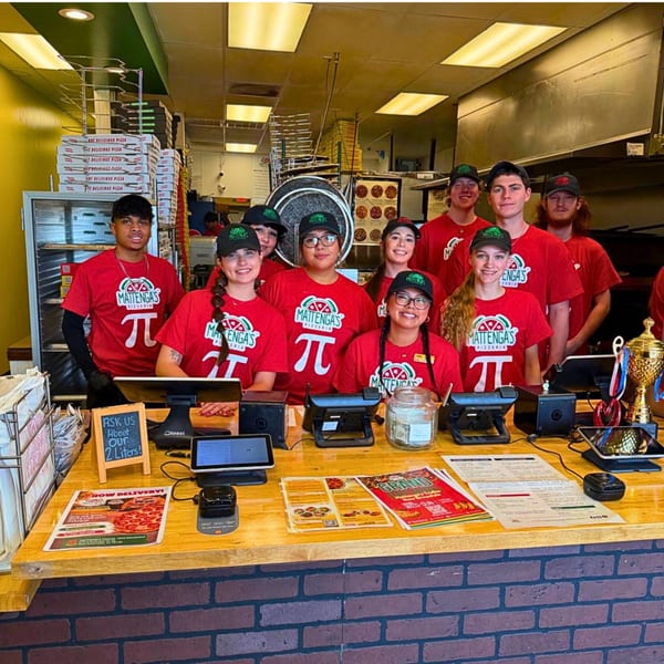 Nine smiling Mattenaga's Pizzeria employees in red shirts and hats stand behind a counter.
