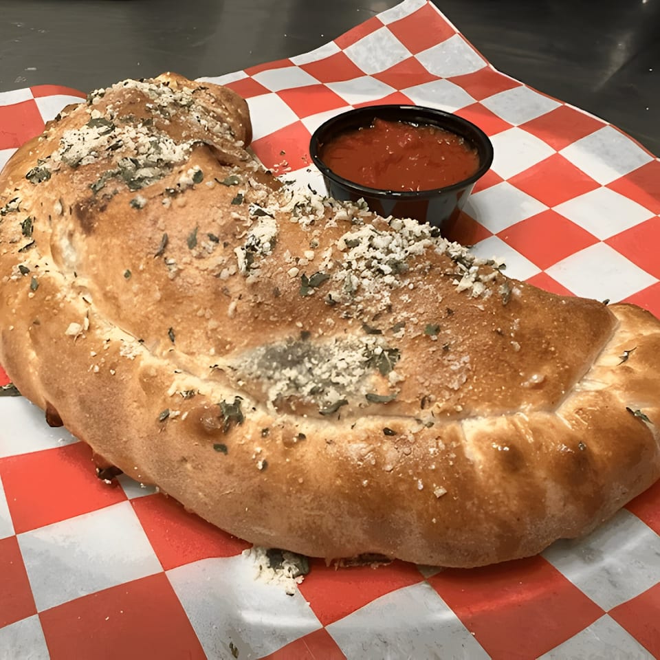 A golden-brown calzone from Mattenga's Pizzeria, topped with herbs and cheese, served with a side of marinara.