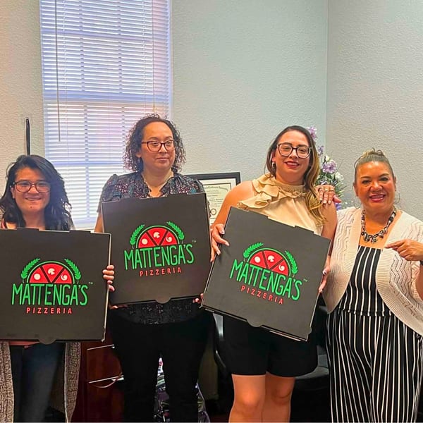 Four women smiling, holding pizza boxes with the Mattega's Pizzeria logo in an office setting.