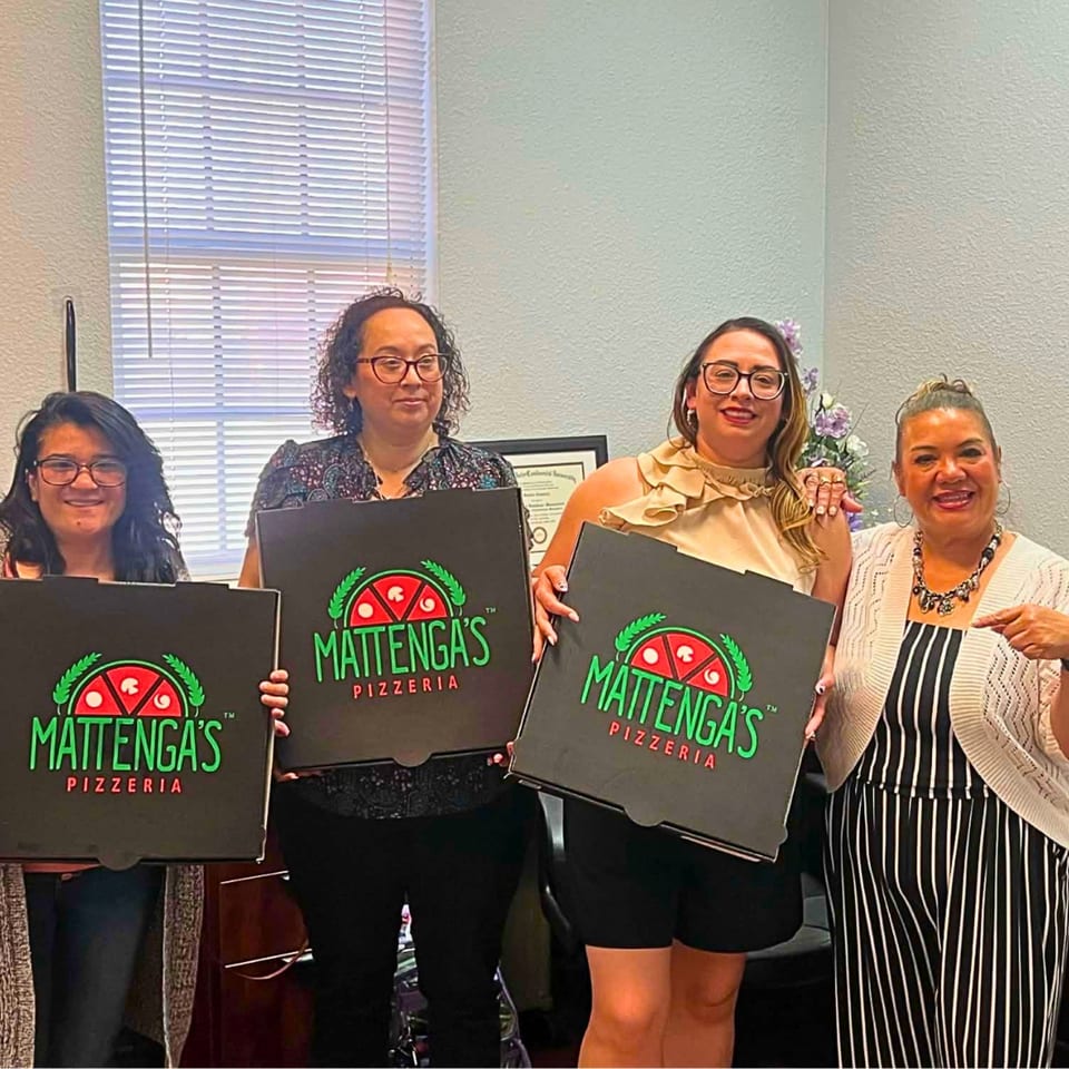 Four women smiling, holding pizza boxes with the Mattega's Pizzeria logo in an office setting.